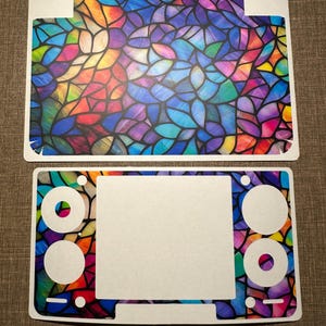 Puede incluir: Juego de pegatinas de dos piezas con un diseño de vidriera. La pieza superior es rectangular con un patrón colorido de azules, morados, verdes y amarillos. La pieza inferior tiene recortes para botones y una pantalla, con el mismo diseño.