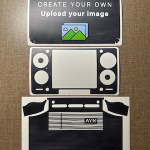 Könnte beinhalten: Drei rechteckige Klebevinyl-Aufkleberbögen für eine tragbare Spielkonsole. Der oberste Bogen zeigt den Text CREATE YOUR OWN Upload your image mit einem Bergsymbol. Der mittlere Bogen ist ein schwarzer Rahmen. Der untere Bogen ist schwarz mit dem Text AYN.