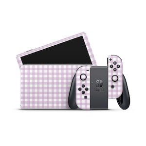 Puede incluir: Una consola Nintendo Switch y sus mandos con un patrón de cuadros vichy lila y blanco. La consola está en una estación de acoplamiento y los mandos están conectados. La pantalla de la Switch es negra.