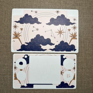 Puede incluir: Dos pegatinas para portátil con un diseño celestial. El diseño incluye nubes azul marino, estrellas doradas y constelaciones sobre un fondo rosa claro. La pieza superior es rectangular y la inferior tiene recortes.