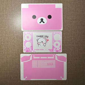 May include: A set of three pink adhesive skins for a handheld gaming console. The top piece features a bear face, the middle piece has a floral pattern with a 'THANK YOU' sticker, and the bottom piece displays the 'AYN' logo and vent cutouts.