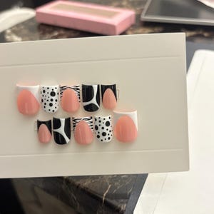May include: A set of artificial nails with various designs. The nails feature a mix of solid peach, black and white patterns, including polka dots, zebra stripes, and geometric shapes. The nails are arranged on a white card.