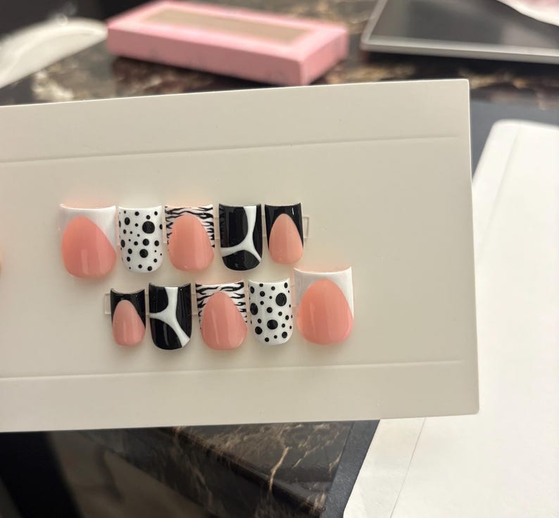 May include: A set of artificial nails with various designs. The nails feature a mix of solid peach, black and white patterns, including polka dots, zebra stripes, and geometric shapes. The nails are arranged on a white card.