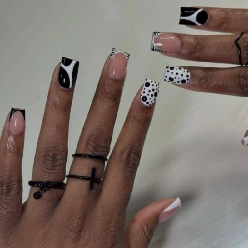 May include: Close-up of a hand with various nail designs. The nails feature black and white patterns, including polka dots, zebra stripes, and abstract shapes. The hand also has black rings with cross and ball details.