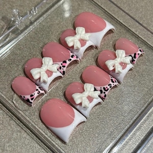 Könnte beinhalten: Set aus ovalen, pinkfarbenen und weißen Kunstnägeln mit weißer Spitze und Leopardenmuster. Jeder Nagel hat oben eine weiße Schleife.