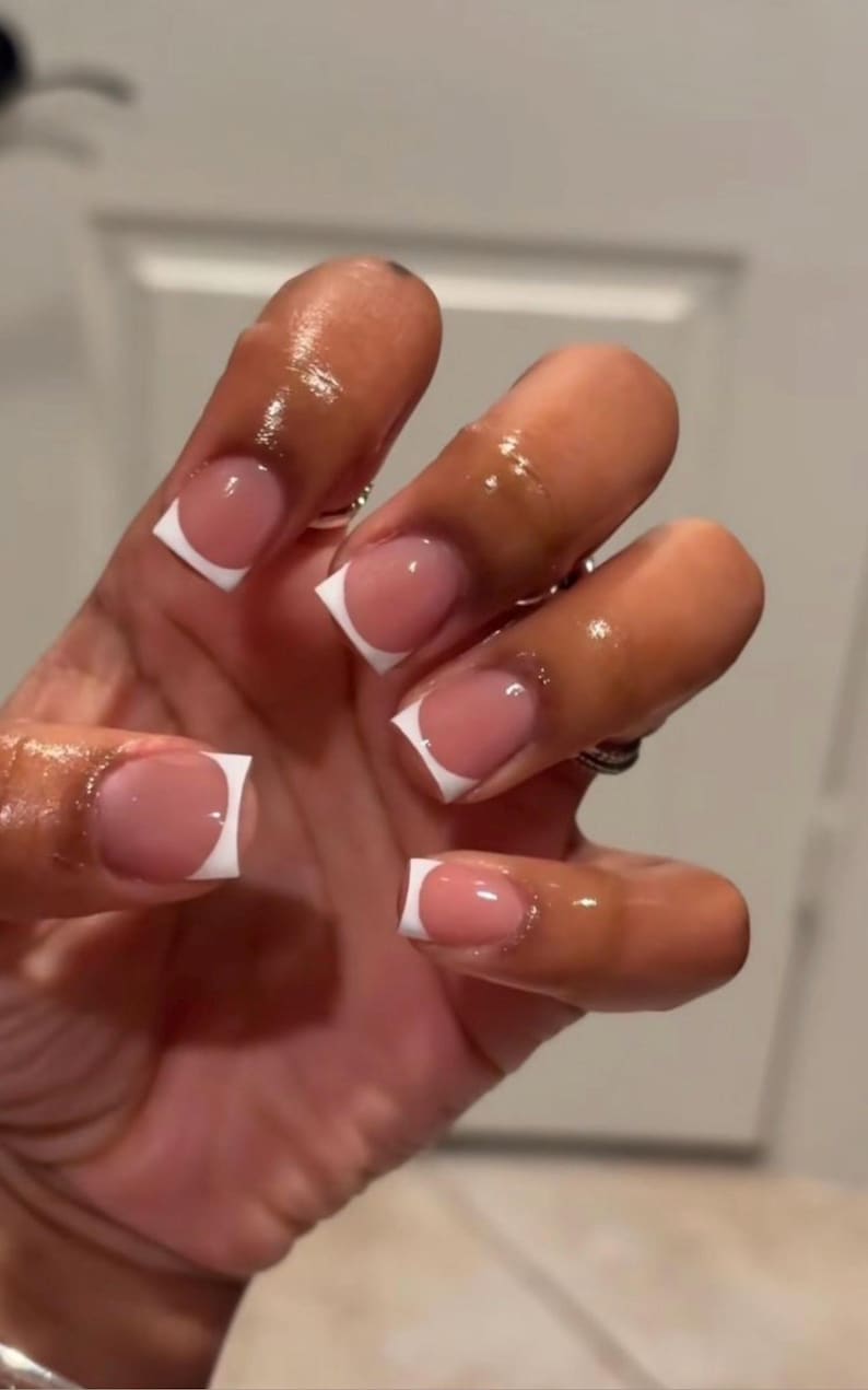 May include: A set of square-shaped artificial nails featuring a classic French manicure design with a glossy, nude-toned base and crisp white tips. The nails are applied to the fingers, which appear coated in a thin layer of oil or moisturizer.