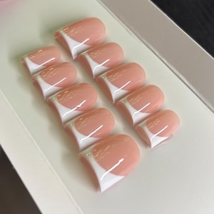 May include: A set of ten press-on nails featuring a classic French manicure design with a soft, translucent pink base and a crisp, opaque white tip. The nails are arranged in two rows on a white display card, showcasing a smooth, glossy finish.
