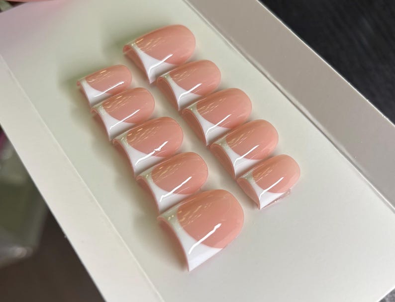 May include: A set of ten press-on nails featuring a classic French manicure design with a soft, translucent pink base and a crisp, opaque white tip. The nails are arranged in two rows on a white display card, showcasing a smooth, glossy finish.