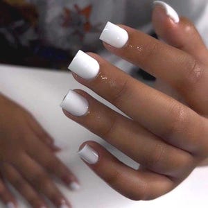 May include: Close-up of a hand with square-shaped, glossy white nail polish. The nails are neatly painted, and the skin tone is a warm brown. The image highlights the manicure's clean lines and bright color.