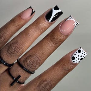 May include: Close-up of a hand with manicured nails. The nails feature a variety of designs, including black and white patterns, a French tip with a black outline, and a polka dot design. The hand also has black rings.