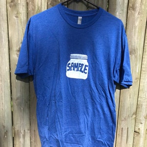Puede incluir: Una camiseta azul con un gráfico blanco de un frasco con la palabra "SAMPLE" en él.