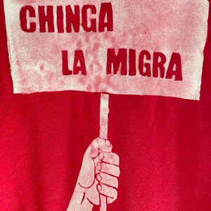 May include: A red background with a white sign held by a hand. The sign reads "CHINGA LA MIGRA" in bold, uppercase letters. The hand and sign are white, contrasting against the red backdrop.