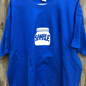 Op de afbeelding: Een koninklijk blauw t-shirt met een witte afbeelding van een pot met het woord "SAMPLE" erop gedrukt.