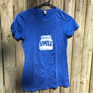 Puede incluir: Una camiseta azul real con un gráfico blanco de un frasco de vidrio con la palabra "SAMPLE" impresa en él.
