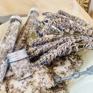 Puede incluir: Tres frascos de vidrio llenos de sales de baño blancas y flores de lavanda secas. Los frascos están sobre una superficie blanca con una cuchara de plata y un ramo de flores de lavanda.