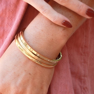 May include: Gold-toned jewelry set featuring three bangles with small diamond accents and a ring with colorful gemstones. A second gold ring with a knot design is also visible. The person is wearing a pink top.
