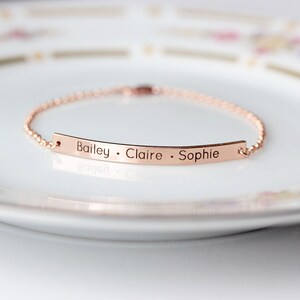 May include: A rose gold chain bracelet with a thin, flat bar charm engraved with the names "Bailey, Claire, Sophie".