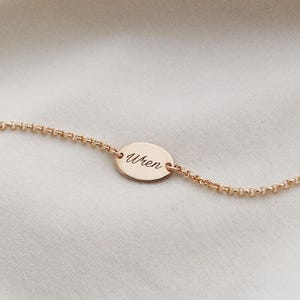 May include: Gold-colored bracelet with an oval pendant. The pendant is engraved with the name "Wren" in a delicate script. The bracelet is set against a soft, neutral background.