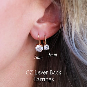 May include: Two gold-tone lever back earrings featuring clear round cubic zirconia stones. One earring measures 7mm and the other 5mm. The text reads: 7mm, 5mm, CZ Lever Back Earrings, LORREN MAE.
