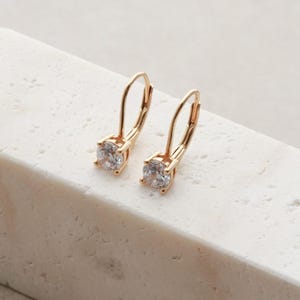 May include: A pair of gold-toned leverback earrings, each featuring a single round, clear, faceted stone set in a four-prong setting. The earrings have a polished metal finish and a classic drop design, showcasing a clear, brilliant-cut gemstone.