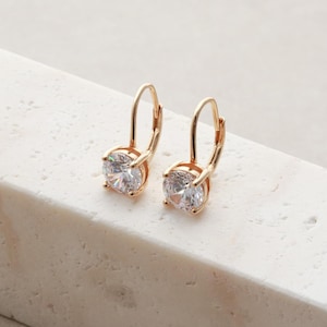 May include: A pair of gold-toned leverback earrings, each featuring a single round-cut clear white gemstone held in a four-prong setting. The earrings have a polished metallic finish and a curved wire closure that secures the stone in place.