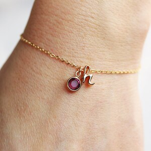 May include: A gold chain bracelet with a small, round, dark purple gemstone and a gold charm with the letter 'h' hanging from it.