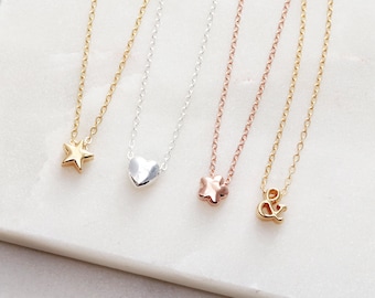 Puede incluir: Cuatro delicados collares de oro con diferentes dijes: una estrella, un corazón, una flor y un símbolo de ampersand. Los collares se muestran sobre una superficie de mármol blanco.