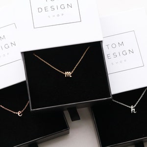 May include: Three small boxes with black interiors, each containing a delicate gold chain necklace with a small letter pendant. The boxes are white with the text "TOM DESIGN SHOP" printed on the front.