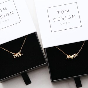 May include: Two gold necklaces in black boxes. The first necklace has the letters "H & K" intertwined. The second necklace has the letters "J & M" with a small heart between the letters. Both boxes have the text "TOM DESIGN SHOP" on the top.