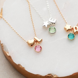 May include: Three gold necklaces with charms and gemstones. The first necklace has a star and the letter "E" charm with a green gemstone. The second necklace has a star and the letters "A" and "O" charms with a blue gemstone. The third necklace has a heart and the letter "L" charm with a pink gemstone.