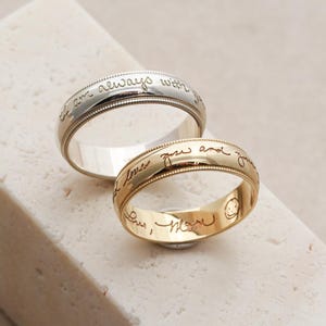 Könnte beinhalten: Zwei Metallringe mit Perlenkanten. Der silberfarbene Ring ist mit dem Text "I am always with you" graviert. Der goldfarbene Ring trägt außen die Gravur "I love you and you" und innen "Love, Mom" zusammen mit einem lächelnden Gesicht.