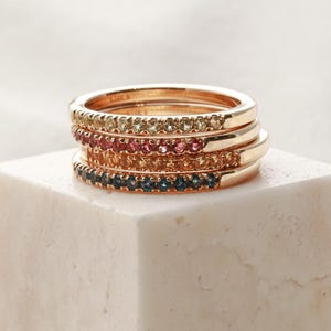 May include: Four gold-colored rings stacked on a white cube. Each ring is adorned with a row of small, colorful gemstones in shades of yellow, red, orange, and blue. The rings are displayed against a soft, white background.