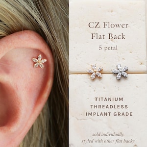 May include: A five-petal flower stud earring featuring clear cubic zirconia stones. The earring is threadless and made of implant-grade titanium. Text on the display reads: CZ Flower Flat Back 5 petal, TITANIUM THREADLESS IMPLANT GRADE.