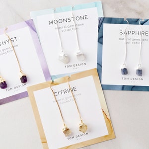 May include: Four cards with different gemstone earrings. Each card has a gold or silver chain with a gemstone at the end. The cards are labeled "Amethyst", "Moonstone", "Sapphire", and "Citrine".