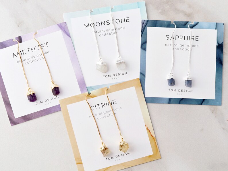 May include: Four cards with different gemstone earrings. Each card has a gold or silver chain with a gemstone at the end. The cards are labeled "Amethyst", "Moonstone", "Sapphire", and "Citrine".