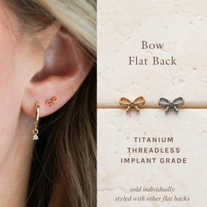 May include: Gold and silver bow-shaped earrings with flat backs. The earrings are displayed next to the text "Bow Flat Back" and "Titanium Threadless Implant Grade". One earring is worn on an ear. The earrings are sold individually.