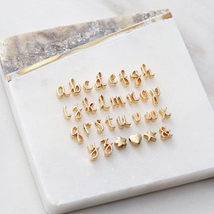 May include: A set of gold-colored letter charms, including the alphabet, a star, a heart, a cloud, and an ampersand. The charms are arranged on a white marble surface.