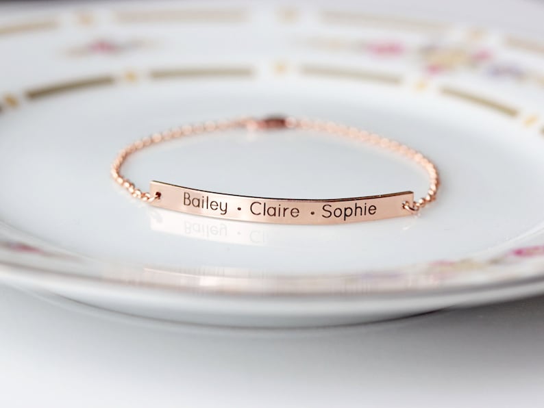 May include: A rose gold chain bracelet with a thin, flat bar charm engraved with the names "Bailey, Claire, Sophie".