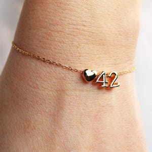 May include: A gold-toned chain bracelet featuring a small, polished heart charm positioned next to the number 42. The bracelet is constructed with a delicate link chain and includes the numerals 4 and 2 as distinct, metallic gold-colored charms.