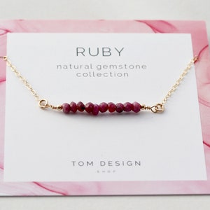 May include: A delicate gold chain necklace with a row of small, deep red ruby gemstones. The necklace is displayed on a pink card with the text "RUBY natural gemstone collection TOM DESIGN SHOP".