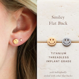 May include: Gold and silver smiley face flat back earrings. The earrings are made of titanium and are threadless. The image also shows a close-up of an ear with a gold smiley face earring and a small stud.