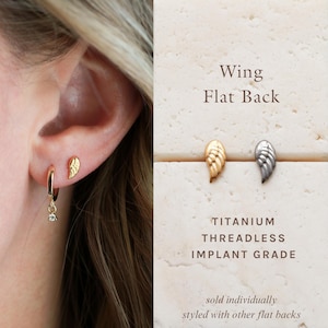 May include: Gold and silver-colored wing-shaped earrings. The earrings are displayed on a cream-colored surface. The text on the image reads "Wing Flat Back" and "Titanium Threadless Implant Grade". One earring is worn on an ear.