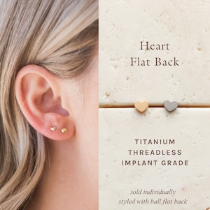 May include: A close-up of a person's ear with a small gold heart stud earring. The image also shows three heart-shaped earrings in gold, silver, and black, with the text "Heart Flat Back" and "Titanium Threadless Implant Grade" below. The text "sold individually styled with ball, flat back" is at the bottom of the image.