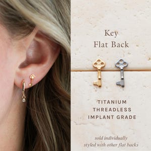 May include: Gold and silver key-shaped earrings. One gold key earring is worn in the ear, and a gold hoop earring with a dangling charm is also worn. The image also shows two key earrings, one gold and one silver, with the text "Key Flat Back".