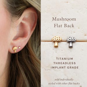 May include: Gold-colored mushroom stud earrings and a small clear stud are shown on an ear. Two mushroom earrings are displayed, one gold and one silver. The text reads "Mushroom Flat Back" and "Titanium Threadless Implant Grade."