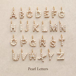May include: A collection of gold-colored alphabet letter pendants, each encrusted with small, round pearls. The letters A through Z are displayed, each with a small loop at the top for hanging. The text "Pearl Letters" is at the bottom.