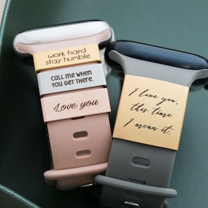 May include: Two silicone watch bands, one blush pink and one dark gray, each featuring metallic engraved charms. The pink band has three charms reading "work hard stay humble," "CALL ME WHEN YOU GET THERE," and "Love you." The gray band has one charm reading "I love you, this time I mean it."