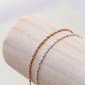 May include: Two delicate chains, one gold and one silver, are displayed on a wooden surface. The chains are both rope style and are a popular choice for necklaces or bracelets.