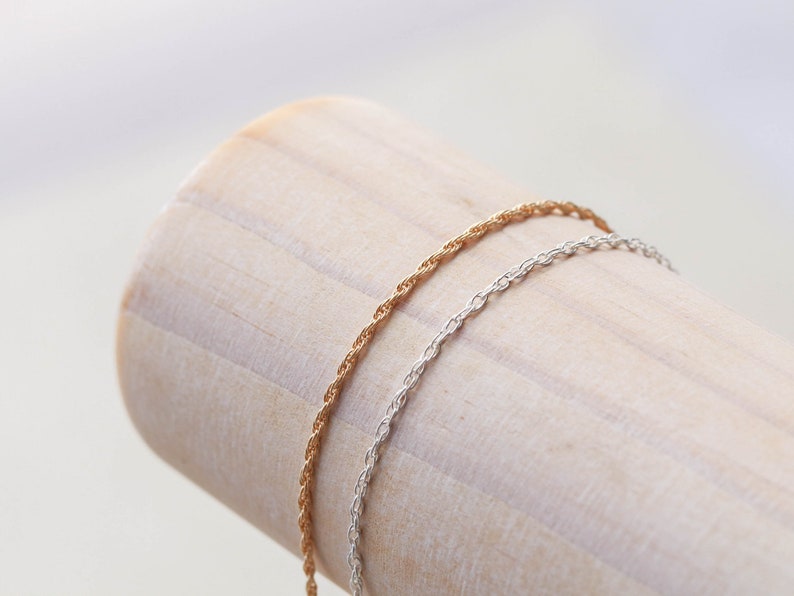 May include: Two delicate chains, one gold and one silver, are displayed on a wooden surface. The chains are both rope style and are a popular choice for necklaces or bracelets.