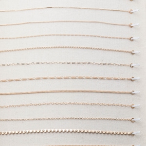 May include: A display of 14 different gold chains, each with a unique link style, hanging from white hooks on a beige fabric background.